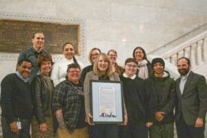 Group of Avivo staff, an Avivo program participant, Wildflyer Coffee staff, and City of Minneapolis staff pose for a photo at Minneapolis City Hall.